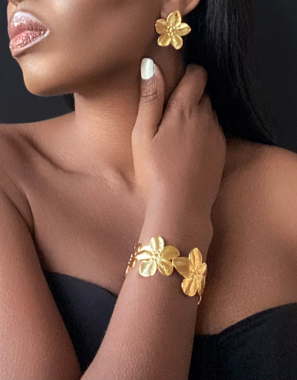 Elegant close-up showcasing a model wearing matching gold flower-shaped earrings and a floral bangle bracelet. The jewellery's intricate design and polished finish exude sophistication, perfect for elevating any outfit. The black backdrop highlights the luxurious gold tones, emphasizing the timeless beauty of these statement accessories.