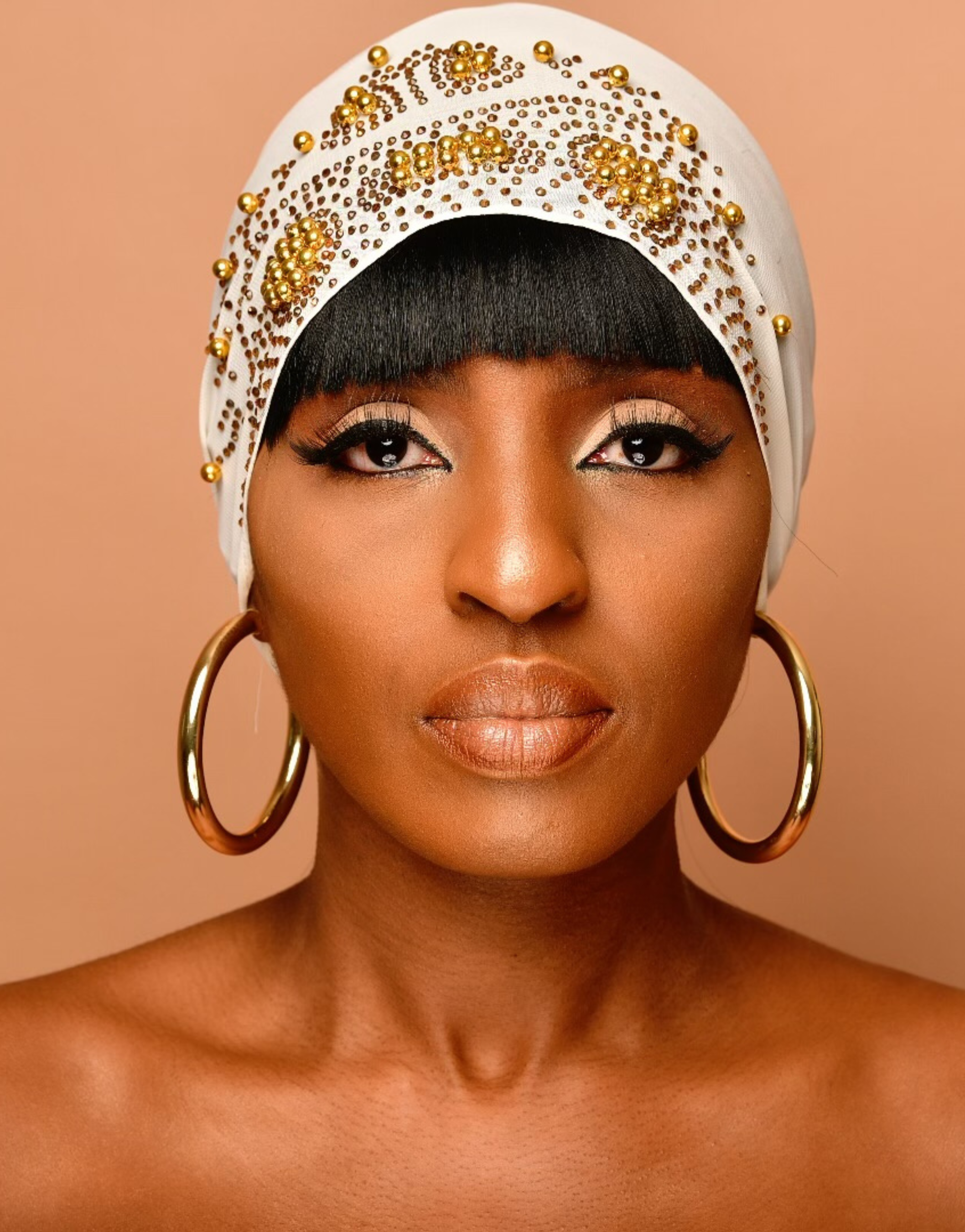 Portrait of a confident woman wearing CAIRO Gold Hoop Earrings and a decorative white headscarf with gold bead detailing. The large, circular gold earrings complement her striking look against a soft peach background.