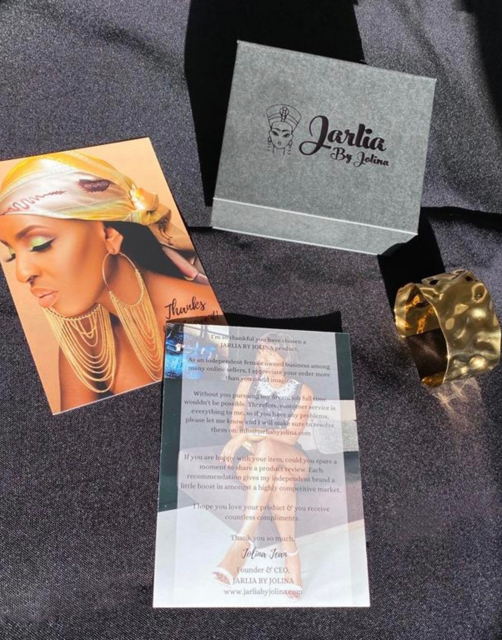 Marketing display for Jarlia By Jolina showcasing a branded gift box with the logo prominently displayed, a hammered gold cuff bracelet, a promotional flyer featuring a model adorned in Jarlia’s jewelry, and a thank-you card emphasizing the brand’s ethos. The items are elegantly arranged on a black textured surface, highlighting the luxurious appeal of the brand.