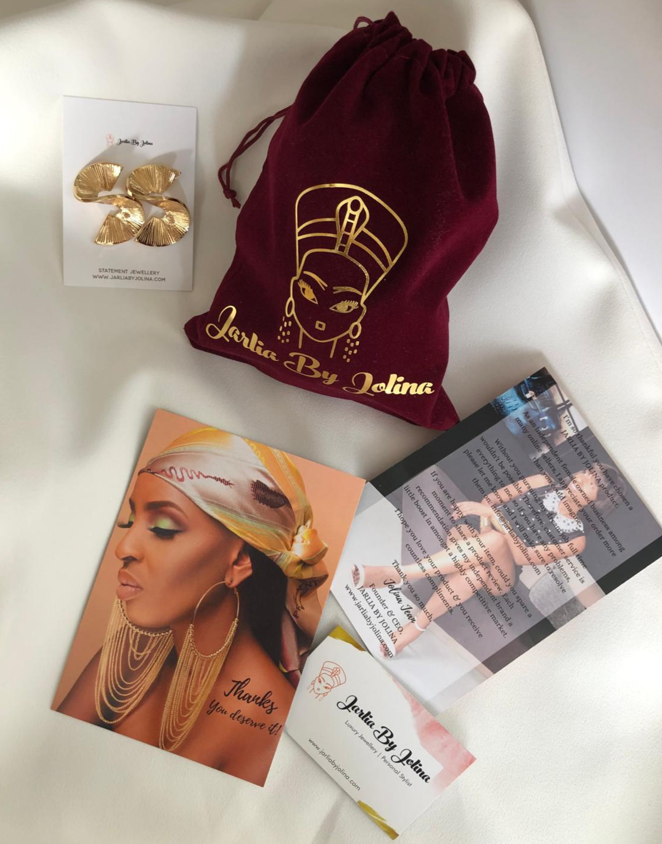 Marketing materials layout for Jarlia By Jolina featuring gold fan-shaped earrings on a branded card, a dark red velvet pouch with gold logo, and promotional flyers with images of a model adorned in Jarlia's jewelry, all arranged neatly on a white surface.